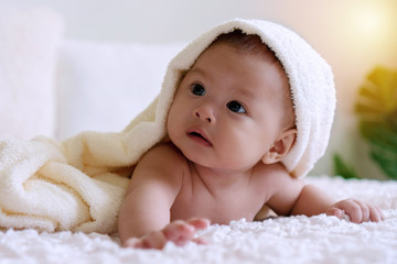 Cheerful cute baby lying under white blanket looking at something. Innocence baby crawling on white bed with towel on his head at home.