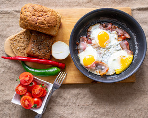 fried eggs with bacon in a pan. near cherry tomatoes chilli peppers rustic bread.