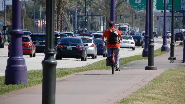Cyclist Cycling Down Sidewalk In Hot Summer Heat With Lots Of Traffic And Heat Waves With Disney Epcot Sign In Distance