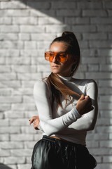 Fashionable brunette girl in a white sweater, orange glasses and black leather pants posing on an white bricks background.