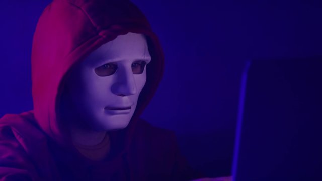 Young anonymous hacker man in red hoodie and mask working on the laptop in the room filled with smoke. Close up slow motion shot