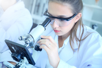 Young female scientist looking through microscope, running an experiment or research. Light blue tone image.
