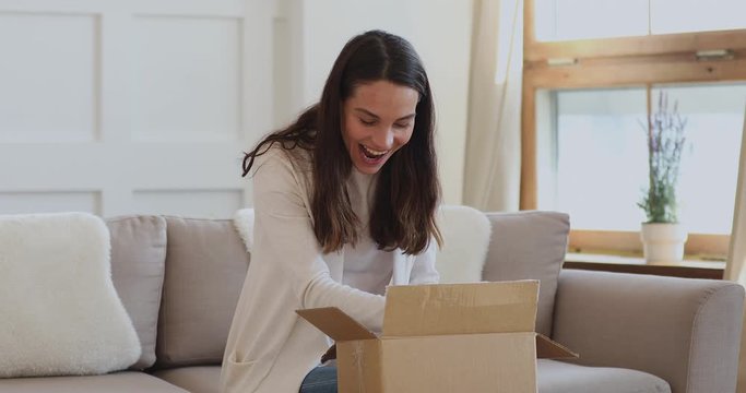 Excited young woman customer opening parcel box at home. Amazed happy girl shopper unboxing fashion purchase sitting on couch. Satisfied female consumer unpacking postal shipping delivery concept