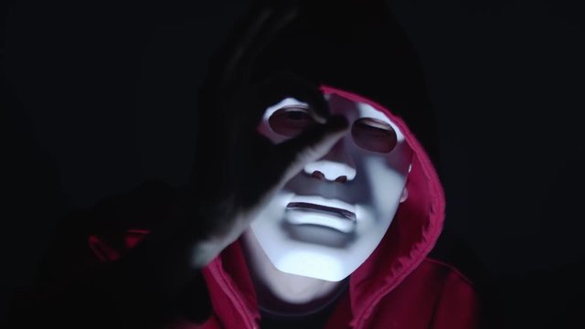 Ok hand gesture or i see you message. Hacker with mask in red hoodie in the dark room illuminated by flash light and surrounded by smoke