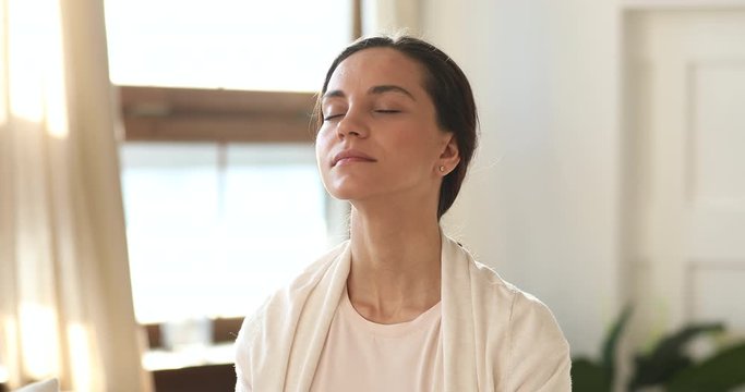 Serene mindful young adult woman meditating with eyes closed at home. Calm millennial girl doing yoga exercise breathing fresh air feeling balance, peace of mind in cozy apartment. No stress concept