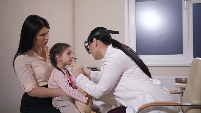 Cute little girl is afraid during examination by woman otolaryngologist. Preadolescent child sitting on mom's knees and going to cry while asian physician trying to examine her nose in hospital ward