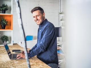 happy smiling remote online working man with laptop, mobile phone and notebook in casual outfit standing up in front of a work desk in an coworking office