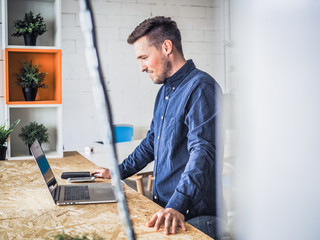 happy smiling remote online working man with laptop, mobile phone and notebook in casual outfit standing up in front of a work desk in an coworking office