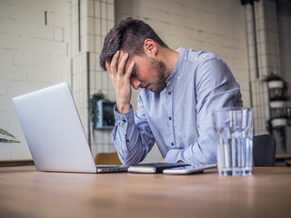 worried and disappointed remote online working man in casual outfit with laptop sitting in an coworking / home office at a work desk - obrazy, fototapety, plakaty