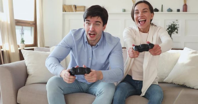 Happy excited man and woman friends gamers holding gamepads playing video game together. Funny young couple winning videogame, having fun, hugging, celebrating victory sitting on couch in living room.