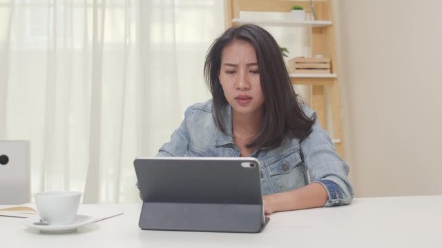 Frustrated Young Asia lady having problem with not working tablet computer sitting on desk. Freelance smart business women casual wear using tablet working in living room at home office. Slow motion.