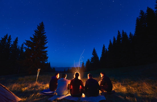Summer Camping Under Stars. Rear View Of Group Of Five Hikers, Men And Woman Sitting Near Bright Bonfire, Tourist Tent Under Dark Night Sky With Sparkling Stars. Concept Of Tourism, Night Camping.