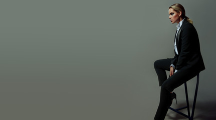 Beautiful sexy slim girl in a men's pantsuit and white shirt is sitting in a studio on a high chair.