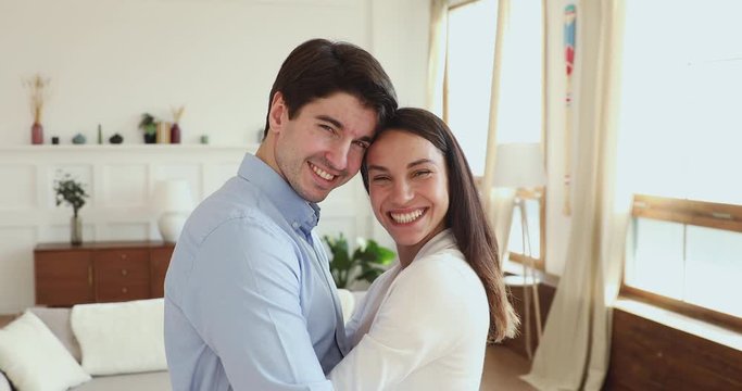 Happy romantic couple bonding embracing looking at camera standing at home. Smiling young husband and wife cuddling, proud millennial first time home owners hugging in new apartment. Portrait.