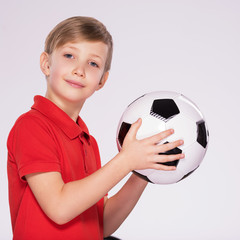 Pretty 8 years old kid in a red t-shirt with a soccer ball in hand.  Photo of a boy in sportswear holding soccer ball, posing at studio. White child holds a soccer ball.
