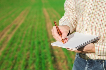 Man in checkered shirt. Farmer hand writing important information in notebook with pen on green...