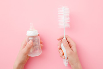 Young mother hands holding baby milk bottle and white brush on pastel pink table background....