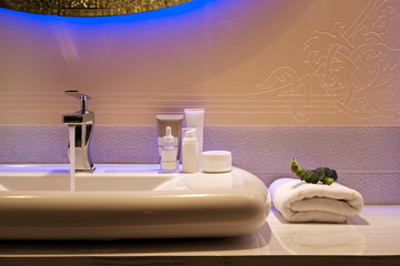 Close up shot of modern interior bathroom sink with blue mirror backlight, liquid soap, cream and other toiletries. White ceramic washbasin in contemporary minimalism style. Copy space, background.