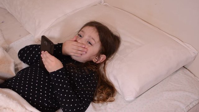 Little charming girl child playing with a mobile phone under the covers on the bed