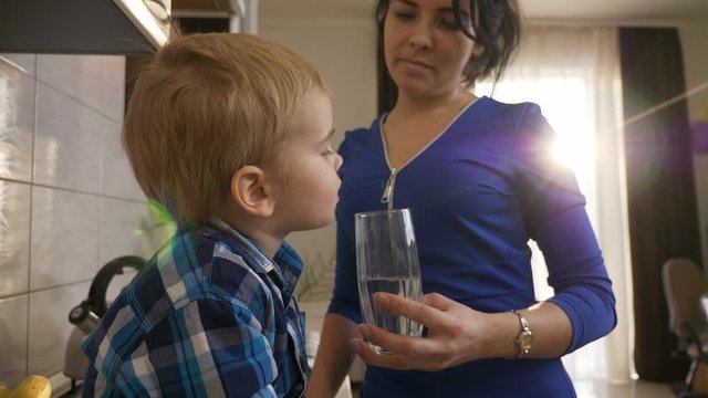 Mother Gives Glass of Water to Son. Little Boy Drinking Water. Child Sitting on Kitchen Countertop. Morning Evening Sunshine through Window. 2x Slow motion, 0.5 speed 60 FPS