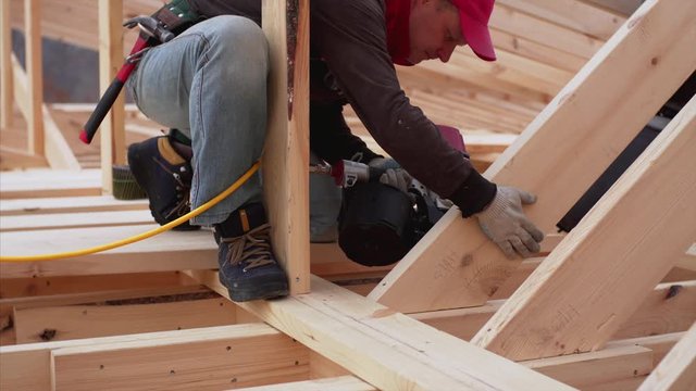 Builder in red cap is nailing wooden planks on the roof with heavy professional pneumatic nail gun. Frame house under construction