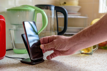 smartphone wireless charging on charging stand on kitchen tabletop. male hand placing phone to...