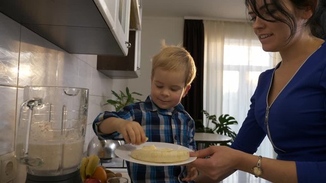 Little Son Helps Mother Cooking Preparing Food for Meal. Child Puts Banana Pieces in Blender Mixer. Child Sitting on Kitchen Countertop. Morning Evening Sunshine through Window. 2x Slow motion, 0.5 sp