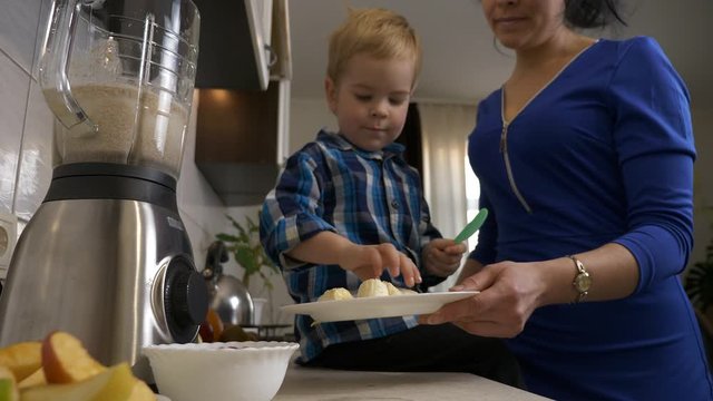 Little Son Helps Mother Cooking Preparing Food for Meal. Child Puts Banana Pieces in Blender Mixer. Child Sitting on Kitchen Countertop. Morning Evening Sunshine through Window. 2x Slow motion, 0.5 sp