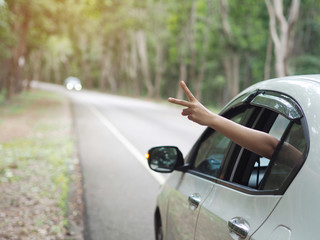 asian woman show her hand out from the window car and show peace sign during road trip on the road. use for travel concept.