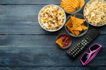 Popcorn, various snacks, 3D glasses and a TV remote control on a blue wooden table. Concept of watching a movie or video at home. top view