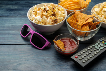 Popcorn, various snacks, 3D glasses and a TV remote control on a blue wooden table. Concept of watching a movie or video at home.