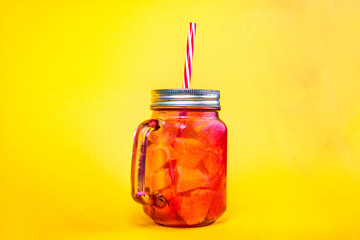 Glass jug with a tin lid and a red straw, with a cold drink made from pieces of fruit.
