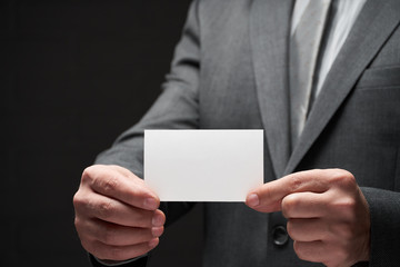 white blank business card closeup in businessman hand, gray suit, dark wall background