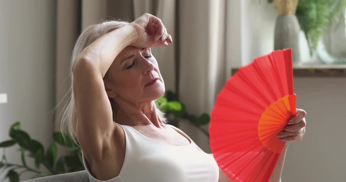 Side view sweaty mature woman sitting on couch, waving paper fan, feeling overheated with no air conditioner at home. Exhausted senior female retiree suffering from hot weather, hormone imbalance.