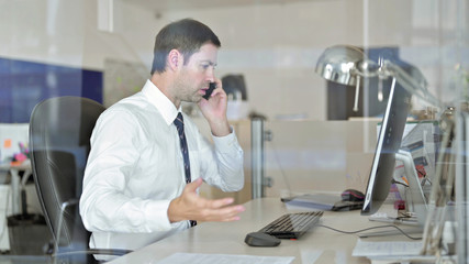 Angry Middle Aged Businessman Talking on his Smartphone and Using Office Computer