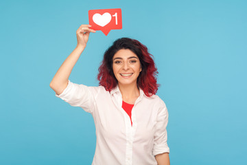 Social media Like button. Portrait of happy woman with fancy red hair holding heart icon over head, recommending to follow and share blog with trendy content. studio shot isolated on blue background