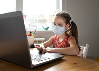 Caucasian preteen girl with medical mask concentrated on her task with laptop. Concept of distance learning in isolation while coronavirus