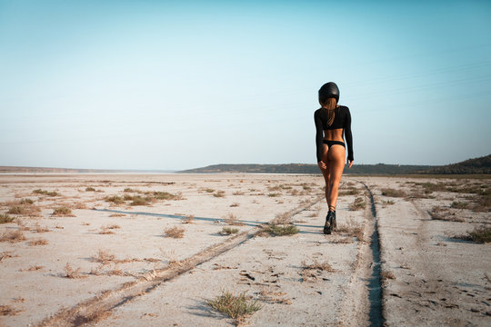 Full length image of Sexy fashion female biker girl in black helmet in dessert. Young beautiful model in black crop top and bikini suit walking away in desert USA. Back view, horizontal, copy space.