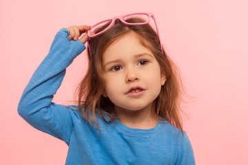 Portrait of amazing beautiful adorable little girl taking off glamour pink glasses and looking with dreamy expression, stylish kid, fashion concept. indoor studio shot isolated on pink background
