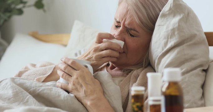 Head shot unhealthy middle aged older woman lying in bed under duvet with hot anti grippe beverage, suffering from runny nose. Unhappy elderly mature granny caught cold flu sneezing, taking medicine.