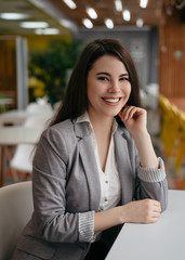 Fototapeta premium Confident businesswoman in modern office, looking at camera, smiling. Portrait of young successful manager sitting at workplace