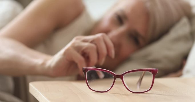 Focus on eyeglasses lying on bedside table. Older mature woman waking up in morning, taking optical glasses from nightstand, close up. Elderly people bad vision, blurred eyesight problem concept.