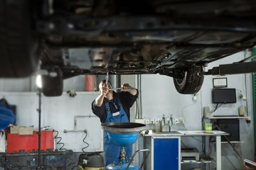 Handsome mechanic in uniform is working in auto service with lifted vehicle. Car repair and maintenance. Oil change