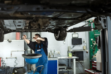 Handsome mechanic in uniform is working in auto service with lifted vehicle. Car repair and maintenance. Oil change