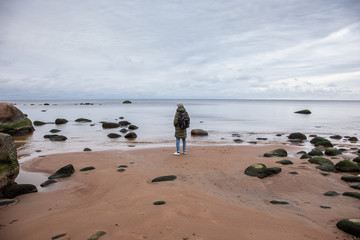 man in warm clothes with a backpack stands on a rocky seashore with his back to the camera