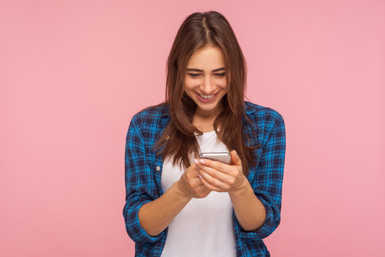 Mobile connection. Portrait of happy brunette girl in checkered shirt reading funny post on social network using cell phone, chatting, typing message. indoor studio shot isolated on pink background