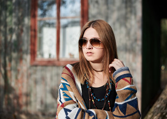 Young blond woman, wearing colorful brown cardigan and sunglasses, posing in front of old time wooden hut in village in autumn. Pretty hippie girl outdoors on weekend, thinking. Three-quarter portrait