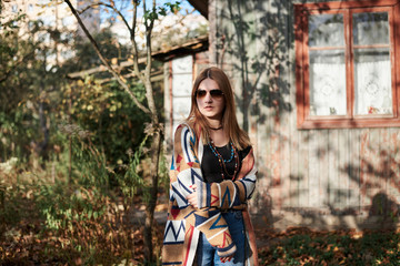 Young blond woman, wearing colorful brown cardigan, posing in front of old time wooden grey hut in village in autumn. Pretty hippie girl having fun outdoors on weekend. Three-quarter portrait.