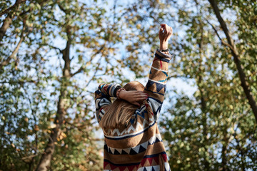 Young blond woman, wearing colorful cardigan, stretching her arms up to the sun in park in autumn. Close-up picture of girl from the back in front of blue sunny sky and green yellow trees in forest.