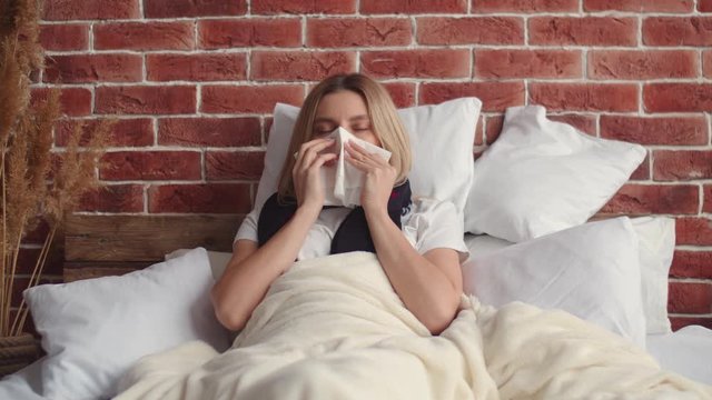 A young woman lies in bed covered with a blanket, coughs covering her mouth with a handkerchief and puts on a medical mask